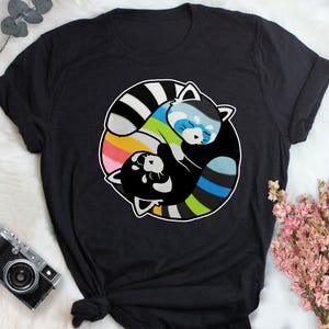 Puede incluir: Camiseta negra con un diseño circular de dos pandas rojos. Uno es blanco y negro, el otro azul y blanco, con un fondo de arcoíris. El diseño está centrado en la camiseta.