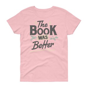 Pode incluir: Camiseta rosa para mulher com o texto "The Book Was Better" em preto. Um pequeno drag&atilde;o verde e um unic&oacute;rnio branco est&atilde;o acima do texto.