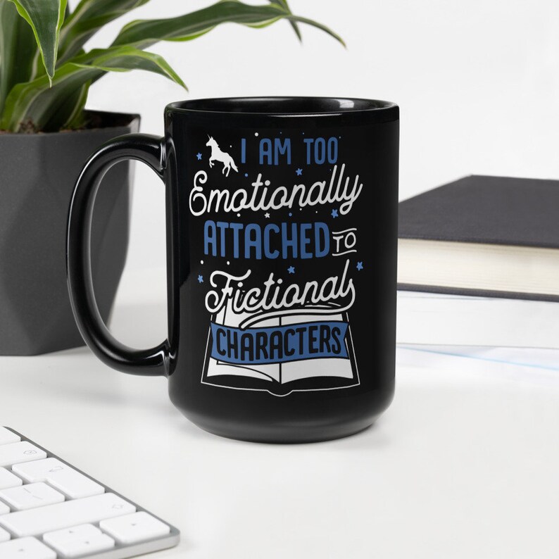 May include: Black coffee mug with a white unicorn and the text "I am too emotionally attached to fictional characters" in blue text. The text is wrapped around a white book with a blue ribbon.