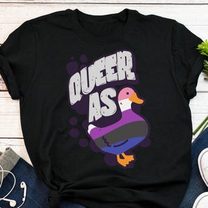 May include: Black t-shirt with the text "Queer as" and a cartoon duck with a purple, black, and blue design.