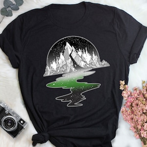 Op de afbeelding: Zwart T-shirt met een witte afbeelding van een bergketen die wordt weerspiegeld in een groene en zwarte rivier. De rivier is gevuld met witte sterren.
