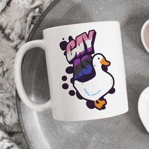 May include: White ceramic mug with a black and white illustration of a goose with the text "Gay as" in rainbow colors.