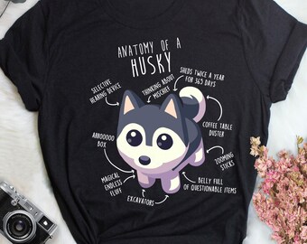 Puede incluir: Una camiseta negra con una ilustración de husky de dibujos animados y el texto "Anatomy of a Husky". La ilustración muestra al husky con varias etiquetas, incluyendo "Dispositivo de audición selectivo", "Pensando en travesuras", "Pelusa mágica sin fin", "Caja Arrooooo", "Excavadora", "Barriga llena de artículos cuestionables", "Plumero de mesa de café", "Palos de zoom" y "Se muda dos veces al año durante 365 días".