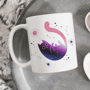 Puede incluir: Taza de cerámica blanca con un diseño de temática galáctica que presenta un gato rosa y morado con estrellas y constelaciones.