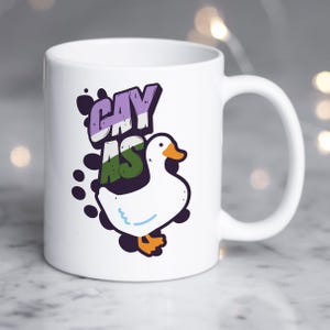May include: White ceramic mug with a cartoon goose and the text "gay as" in a purple, green, and white color scheme.
