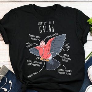 May include: Black t-shirt featuring a detailed diagram of a Galah, labeled "Anatomy of a Galah." The diagram includes humorous labels like "Face Scissors" and "Decibel-Destroying Alert Megaphone," with a pink and gray bird illustration.