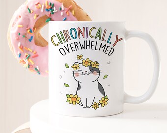 May include: White ceramic mug with a black and white cartoon cat wearing a flower crown. The mug has the text "Chronically Overwhelmed" in rainbow letters.