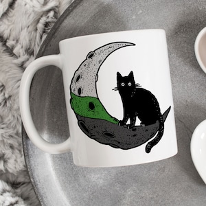 Pode incluir: Caneca de cerâmica branca com um gato preto sentado em uma lua crescente. A lua é cinza, verde e branca com uma textura pontilhada.