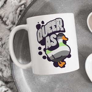 May include: White ceramic mug with a black and white graphic of a duck with a green, gray, and black striped pattern. The text "Queer as" is in a black and white comic book style font.