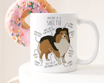 Op de afbeelding: Witte keramische mok met een cartoonillustratie van een Shetland Sheepdog. De illustratie is voorzien van humoristische beschrijvingen van de kenmerken van de hond, zoals "onverdraaglijk pluche kont", "magische eindeloze pluche" en "verliest twee keer per jaar 365 dagen lang haar".