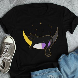May include: Black t-shirt featuring a black cat sleeping on a crescent moon with yellow, purple and white stripes. The moon is surrounded by small stars.
