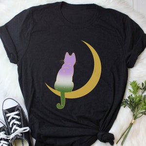 Può includere: Maglietta nera con un gatto viola, verde e bianco seduto su una mezzaluna dorata.