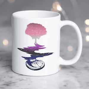 Puede incluir: Taza de cerámica blanca con un diseño de árbol rosa y morado. El árbol se refleja en un charco de agua con una brújula en el fondo.