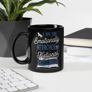 May include: Black coffee mug with a white unicorn and the text "I am too emotionally attached to fictional characters" in blue text. The mug is on a white surface with a keyboard and a stack of books.