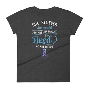 May include: A dark heather grey t-shirt with a blue and white graphic that reads "She believed she could, but she was really tired, so she didn't". A purple awareness ribbon is below the text.