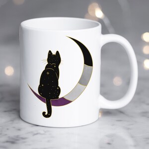 May include: White ceramic mug with a black cat sitting on a crescent moon with purple, gray, and black stripes. The cat is outlined in gold and has gold stars on its body.