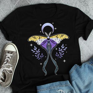 Może przedstawiać: Czarny t-shirt z niebiańskim motywem ćmy. Ćma ma żółte, fioletowe i białe skrzydła, z półksiężycem i gwiazdami nad nią. Dwie gałązki fioletowych liści flankują ćmę. Widoczne są szare trampki i dżinsowe szorty.