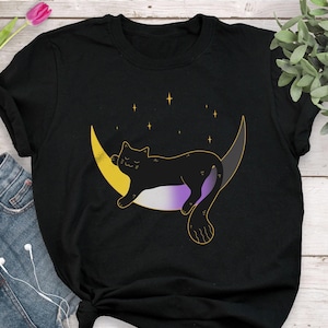 Op de afbeelding: Zwart T-shirt met een zwarte kat die slaapt op een halve maan met gele en paarse accenten. De maan is omringd door sterren.