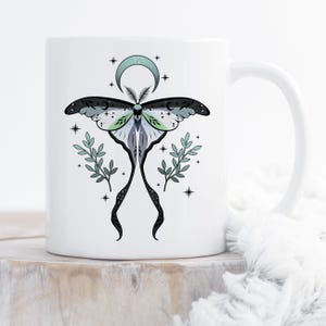 Puede incluir: Taza de cerámica blanca con una ilustración detallada de una polilla, una luna creciente y estrellas. El diseño de la polilla incorpora tonos de negro, gris, verde y blanco, con ramitas de hojas a ambos lados. La taza tiene un asa curva.
