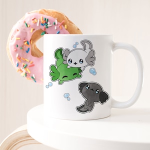 Puede incluir: Taza de cerámica blanca con un diseño de ajolote verde, gris y blanco. Los ajolotes son de estilo de dibujos animados y tienen ojos grandes. La taza tiene un asa negra.