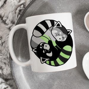 Puede incluir: Taza de cerámica blanca con un diseño de dibujos animados de dos pandas rojos en negro, gris y verde. El diseño está centrado en la taza, con un asa blanca. La taza está sobre una bandeja gris.