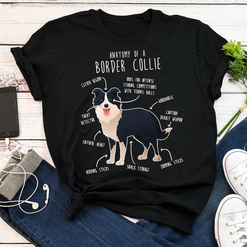 May include: A black t-shirt featuring a white graphic of a border collie dog. The graphic includes text that says "Anatomy of a Border Collie" and lists various traits of the breed, such as "Clever Region", "Treat Detector", "Faithful Heart", "Orbs for Intense Staring Competitions with Tennis Balls", "Workaholic", "Caution: Deadly Weapon", "Herding Sticks", "Snack Storage", and "Zooming Sticks".