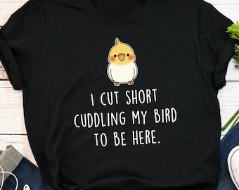 May include: Black t-shirt with a white graphic of a yellow and white bird. The text on the shirt reads "I cut short cuddling my bird to be here."