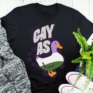 May include: Black t-shirt with the text "gay as" in white and purple lettering. A cartoon duck with a purple head, green, white, and purple feathers, and orange feet is below the text.