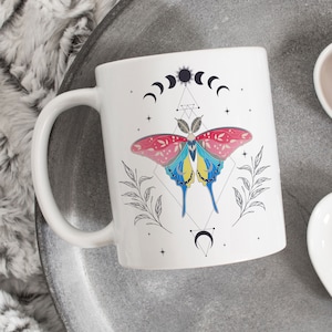 May include: White ceramic mug with a colorful butterfly design, moon phases, and stars. The butterfly is surrounded by geometric shapes and leaves.
