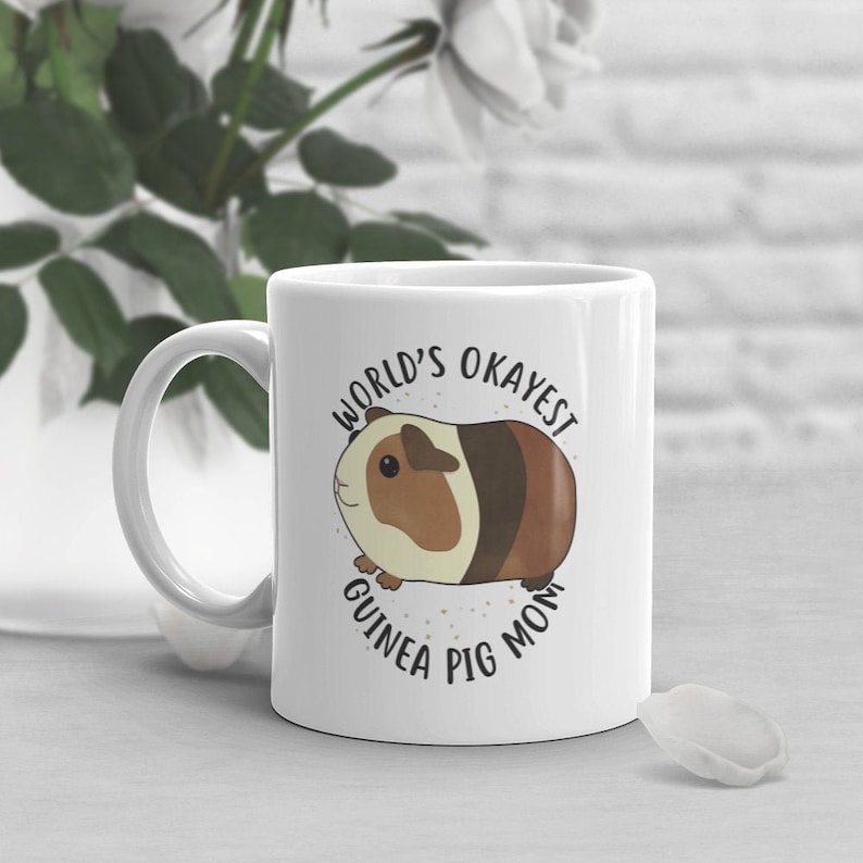 May include: White ceramic mug with a brown and white guinea pig illustration and the text "World's Okayest Guinea Pig Mom".