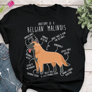 May include: A black t-shirt featuring a white graphic of a Belgian Malinois dog. The graphic is labelled with humorous anatomical features, such as "surveillance system", "sadness detector", "workaholic", and "coffee table duster".