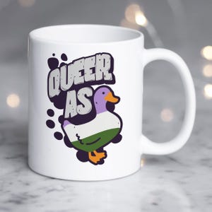May include: White ceramic mug with a cartoon duck in the center. The duck is purple, white, and green and has the words "Queer as" above it.