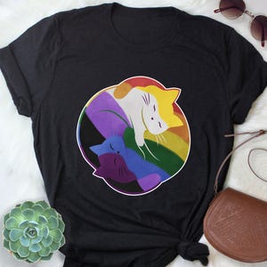 May include: Black t-shirt featuring a colorful graphic of three stylized cats within a rainbow circle. The cats are in shades of yellow, white, purple, blue, and red. The shirt is made of a soft material.