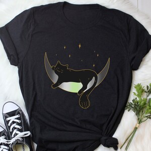 Op de afbeelding: Zwart T-shirt met een zwarte kat die slaapt op een halve maan met een groen, wit en grijs kleurenschema. De maan is omringd door kleine sterren.