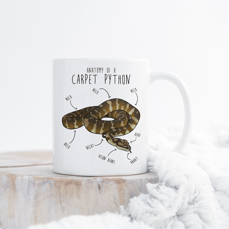 May include: White ceramic mug featuring a cartoon illustration of a brown and tan snake. The mug is labelled "Anatomy of a Carpet Python" and includes labels for the snake's neck, head, and vision beans.