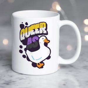 May include: White ceramic mug featuring a cartoon duck with the text "QUEER AS" in a colorful design. The duck has orange feet and beak, with a blue line detail. The mug has a curved handle.