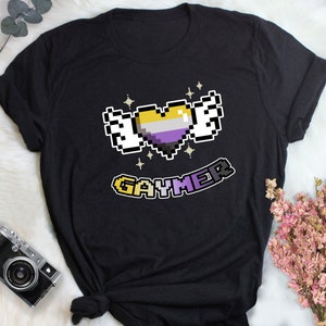 Op de afbeelding: Zwart T-shirt met een gepixelde hart met gele, witte en paarse strepen, vleugels en glitters. De tekst "GAYMER" staat onder het hart in gepixelde letters.