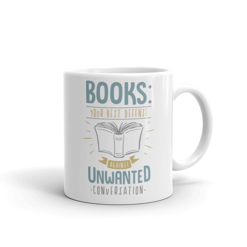 Puede incluir: Taza de cer&aacute;mica blanca con el texto "BOOKS: YOUR BEST DEFENSE AGAINST UNWANTED CONVERSATION" en verde azulado y dorado. El dise&ntilde;o incluye una ilustraci&oacute;n de un libro abierto. Un regalo perfecto para los amantes de los libros.