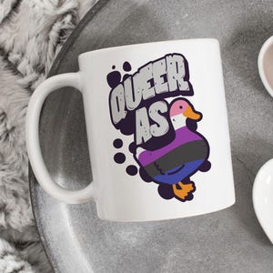 May include: White ceramic mug with a black, purple, and blue duck illustration and the text "Queer as" in a bold, black font.