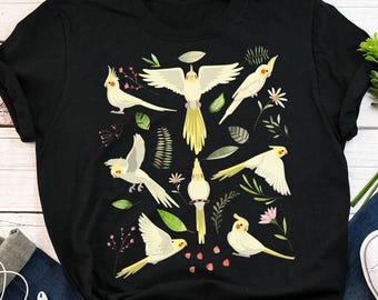 May include: Black t-shirt with a white cockatiel bird design. The design features multiple cockatiels with green leaves and pink flowers.