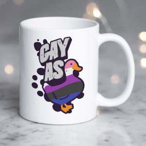 May include: White ceramic mug with a black, purple, and blue duck with the text "gay as" in a gray and black font.