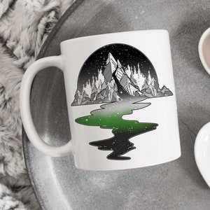 Puede incluir: Taza de cerámica blanca con una ilustración en blanco y negro de una cordillera con un río verde que fluye a través de ella. El río se refleja en el agua de abajo, y el cielo está lleno de estrellas.