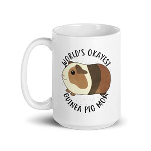 May include: White ceramic mug with a brown and white guinea pig illustration. The text "World's Okayest Guinea Pig Mom" is printed in black around the guinea pig.
