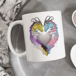 Puede incluir: Taza de cerámica blanca con un diseño de dos dragones formando una forma de corazón. Los dragones son amarillos, azules y morados con un corazón gris en el centro.
