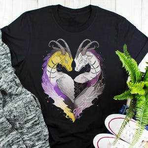 Puede incluir: Camiseta negra con un diseño en forma de corazón de dos dragones. Uno es amarillo, el otro blanco, con detalles morados y grises. Las cabezas de los dragones se tocan, formando un corazón.