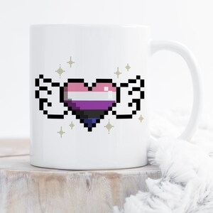 Può includere: Tazza in ceramica bianca con un cuore pixelato con ali, colorato in rosa, bianco, viola, nero e blu. Il disegno è circondato da piccole stelle dorate. La tazza ha un manico curvo.