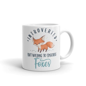 May include: White ceramic mug featuring a cartoon fox illustration and the text "Introverted but willing to discuss foxes".