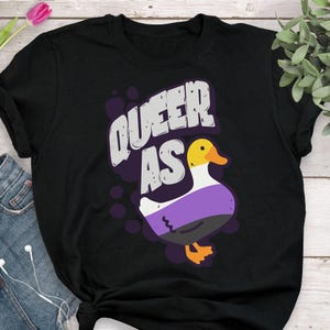 May include: Black t-shirt with the text "Queer as" and a cartoon duck with a purple, white, and black striped pattern.