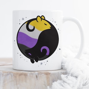 May include: White ceramic mug with a black and white yin yang design featuring two cartoon rats. The yin yang symbol is colored with the colors of the non-binary pride flag: yellow, white, purple, and black.