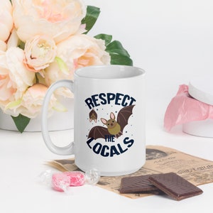 May include: White ceramic mug with a blue and brown graphic of bats and the text "Respect the Locals".